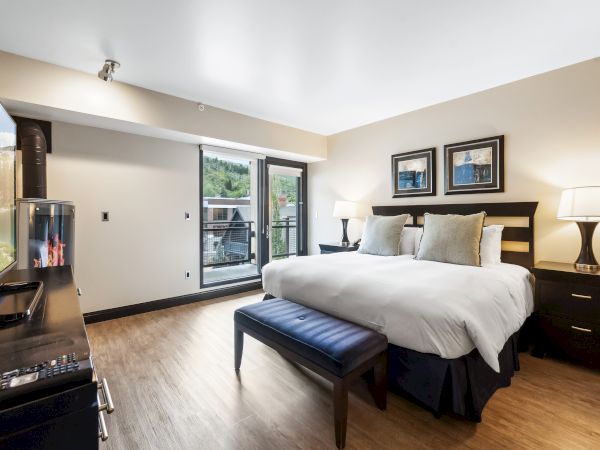 The image shows a modern hotel room with a king-sized bed, flat-screen TV, two lamps, and a balcony view outside the window.