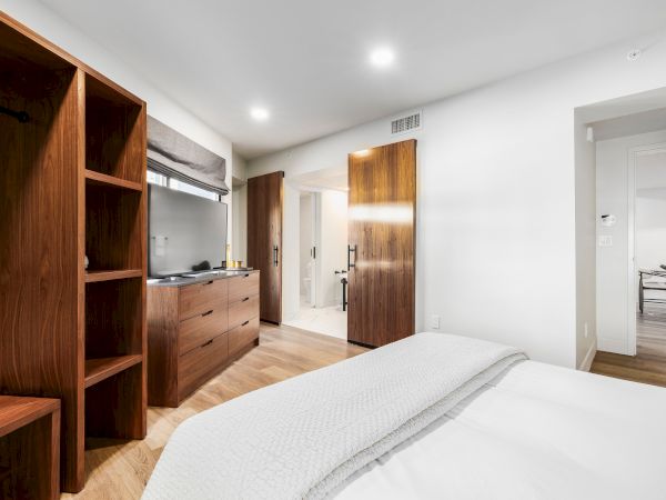 A modern bedroom with a white bed, warm wood built-ins, sleek cabinetry, and an open view to a kitchen/living area.