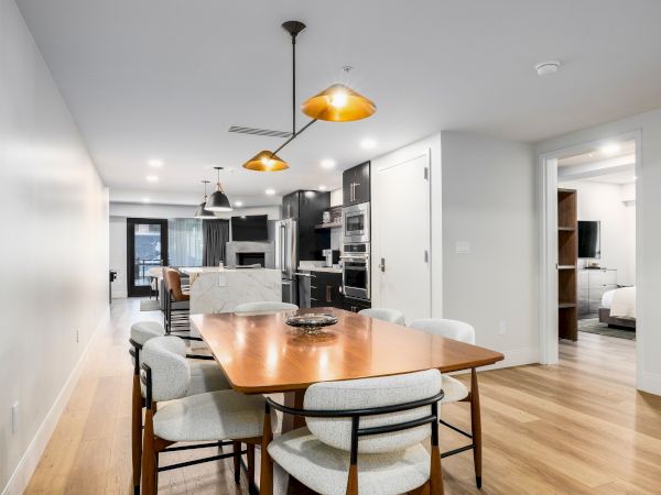 Modern kitchen and dining area with pendant lights, marble island, black cabinets, and a wood table with gray upholstered chairs.