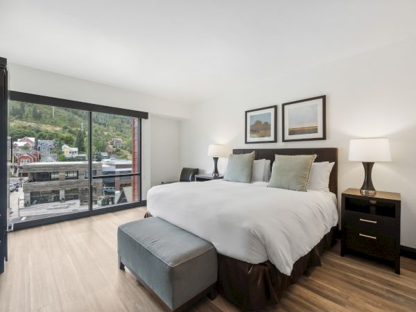 A modern bedroom with a large bed, two lamps, and framed art. A window offers a view of the outdoors with greenery visible.