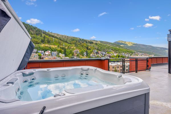 A rooftop hot tub with clear water overlooks a scenic view of hills and houses under a blue sky with a few clouds.