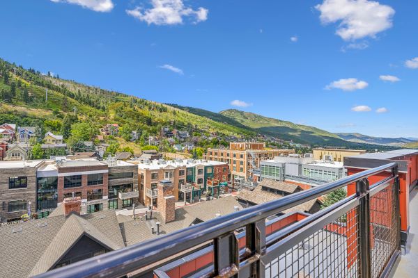 A scenic view from a balcony shows a town with buildings, surrounded by green hills under a clear blue sky with clouds.