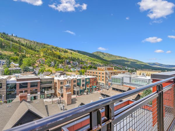 A scenic view from a balcony shows a town with buildings, surrounded by green hills under a clear blue sky with clouds.