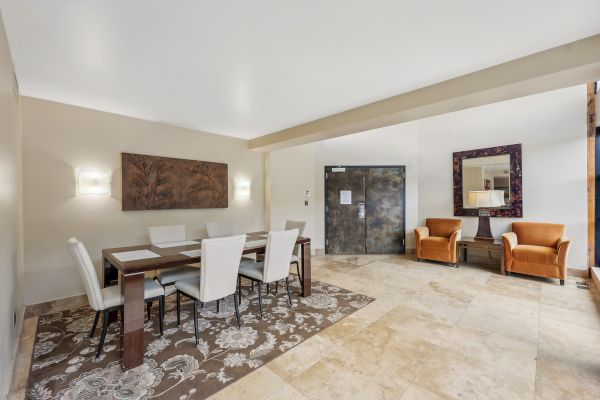 A dining area with a table, six chairs, abstract wall art, two armchairs, a door, a mirror, and a patterned rug on tiled flooring.