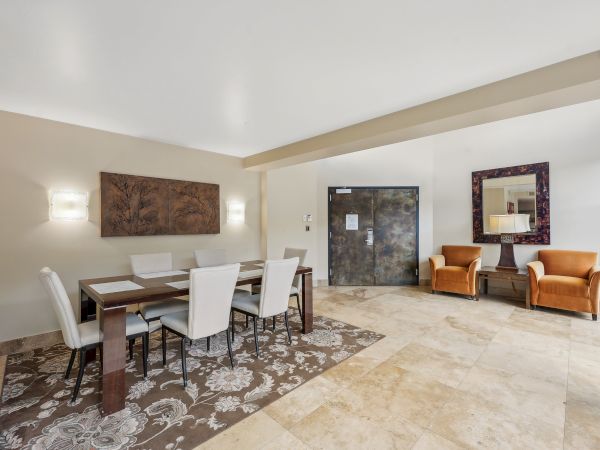 A dining area with a table, six chairs, abstract wall art, two armchairs, a door, a mirror, and a patterned rug on tiled flooring.