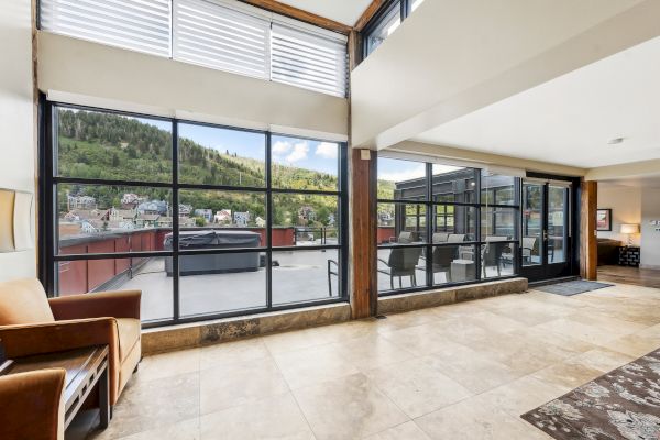 The image shows a modern living space with large windows, a view of a terrace, outdoor seating, and a scenic hillside background.