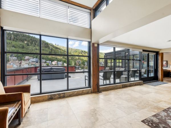 The image shows a modern living space with large windows, a view of a terrace, outdoor seating, and a scenic hillside background.