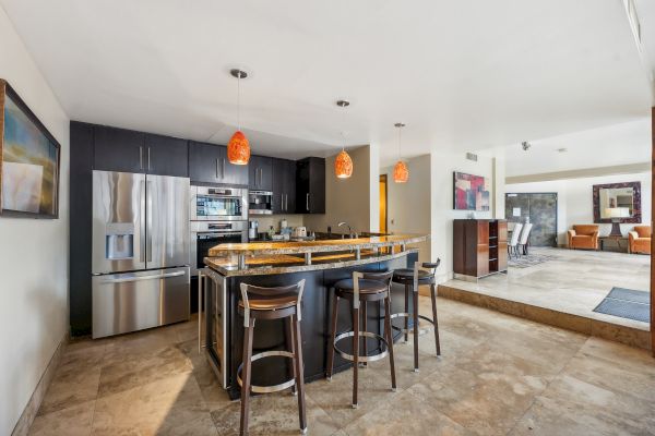 A modern kitchen with an island, bar stools, pendant lights, stainless-steel appliances, and artwork on the walls.