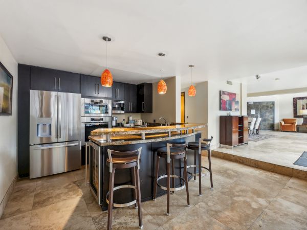 A modern kitchen with an island, bar stools, pendant lights, stainless-steel appliances, and artwork on the walls.