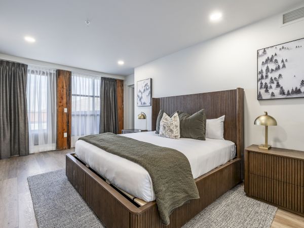 A modern bedroom with a large bed, wood paneling, lamps, framed forest art, and tall curtains by a glass door, styled for comfort.