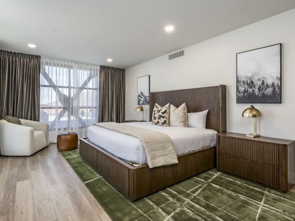 Bright modern bedroom with a large bed, wood headboard, gold lamps, framed art, and curtains.