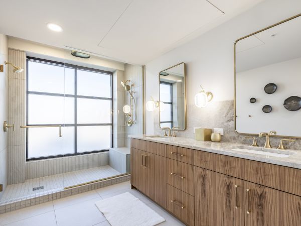 Modern bathroom with glass shower, double vanity, brass fixtures, wall lights, and stone-tile floors.