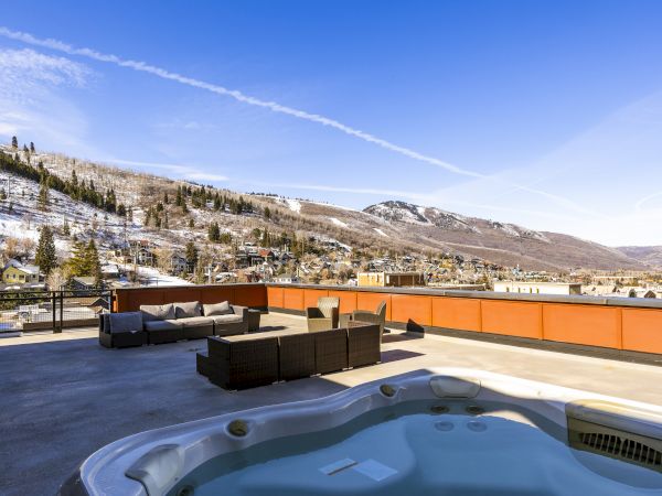 Snowy mountain town view from a sunny patio hot tub with lounge chairs, orange railing, pine trees, and blue sky.
