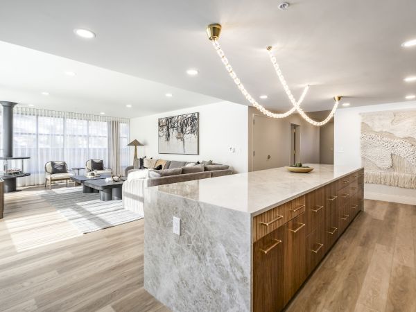 A modern living room and kitchen with a marble island, wood cabinets, sofa seating, large windows, and sparkling pendant lights.
