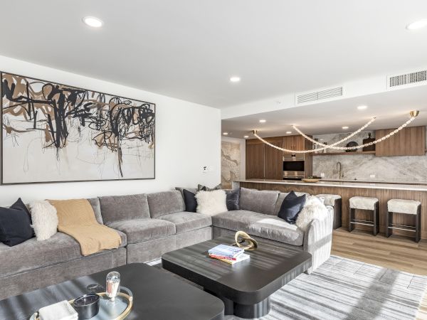 Modern living room with a grey sectional sofa, large abstract wall art, and an open kitchen island with pendant lights.