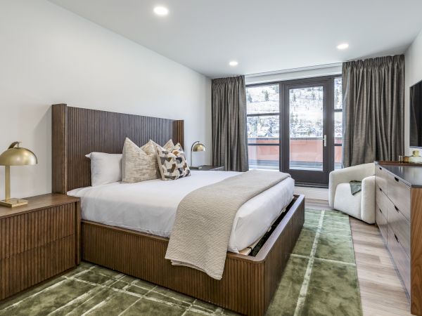 A modern hotel room with a wooden bed, neutral decor, a green rug, bedside lamps, a dresser with a TV, and a balcony view through glass doors.