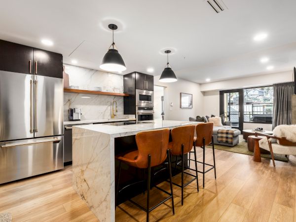 A modern open-plan kitchen and living room with an island, pendant lights, stainless appliances, and cozy seating by the window.