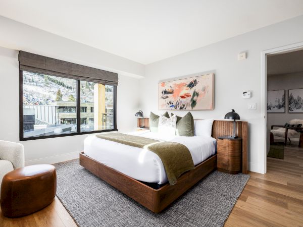 A modern bedroom with a wooden bed, green pillows, large windows, and mountain-themed artwork on white walls.
