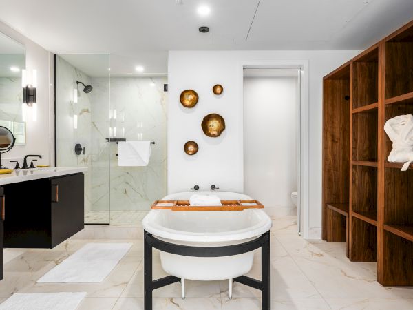 A modern bathroom with a white soaking tub, glass shower, double vanity, gold wall decor, and wooden shelving with towels.