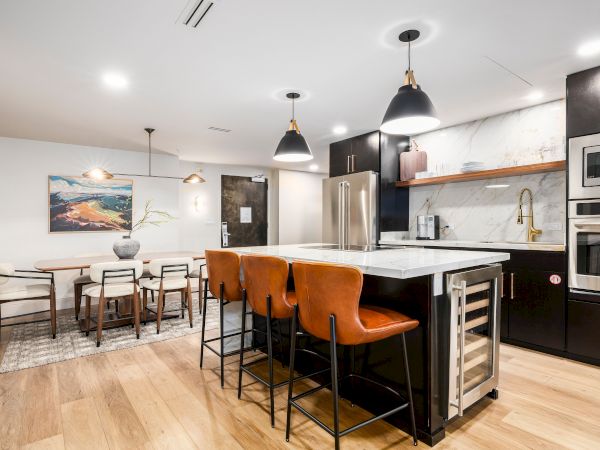 A modern kitchen and dining area with a marble island, stainless appliances, pendant lights, and orange bar stools.
