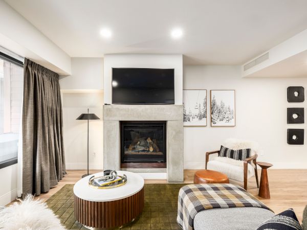 A modern living room with a fireplace, wall-mounted TV, curtains, framed forest art, and seating by a cozy lounge area.