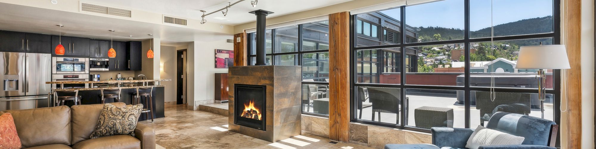 A modern living room with sofa, fireplace, large windows, and a view of a patio and mountains in the background.