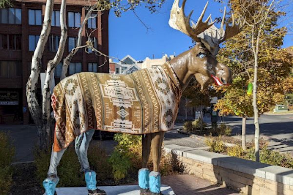 A moose statue dressed in a patterned blanket and blue boot covers stands in a sunny urban setting with trees and buildings nearby.