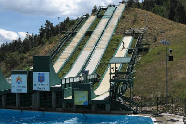 A ski jump training facility with ramps leading into a pool, surrounded by trees and hills under a partly cloudy sky.