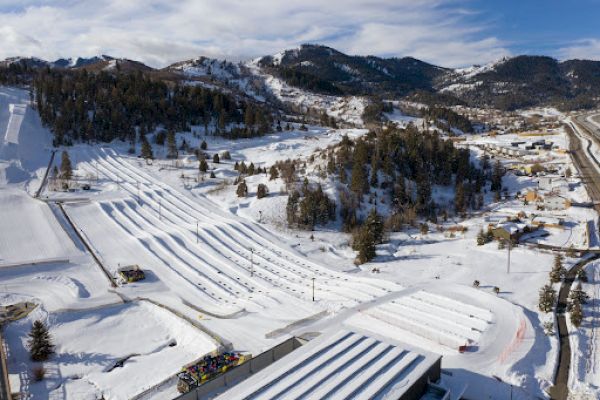 A snowy landscape features ski slopes and surrounding mountains under a partly cloudy sky, with roads and structures visible in the area.