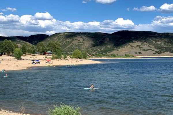 A scenic lake surrounded by hills, with people kayaking and relaxing on a sandy beach under clear blue skies with fluffy clouds.