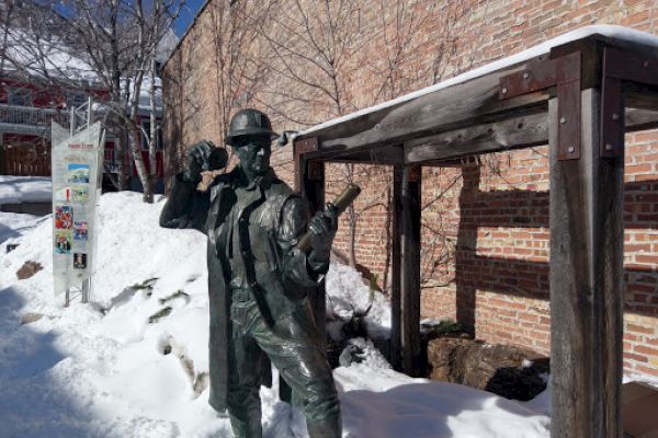 A bronze statue of a miner with tools stands in the snow next to a wooden structure and brick wall.