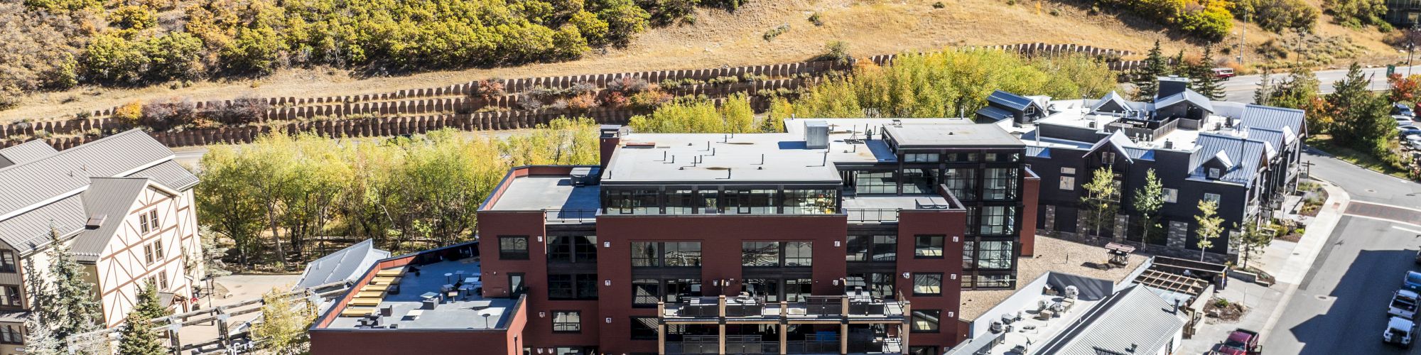 An aerial view of a modern residential complex in front of a hillside with autumn foliage and nearby roads and buildings.