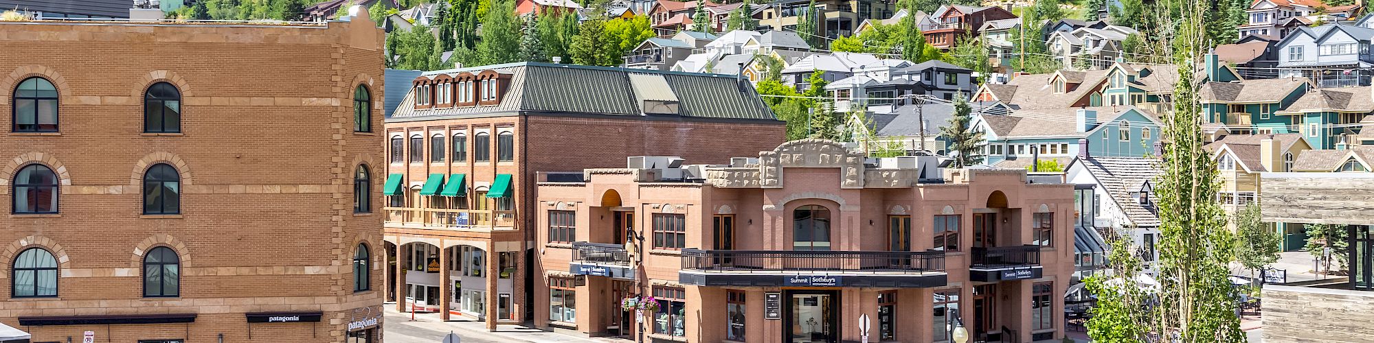 This image shows a quaint, small-town street with brick buildings, nestled in a lush, green mountainous area under a clear blue sky.