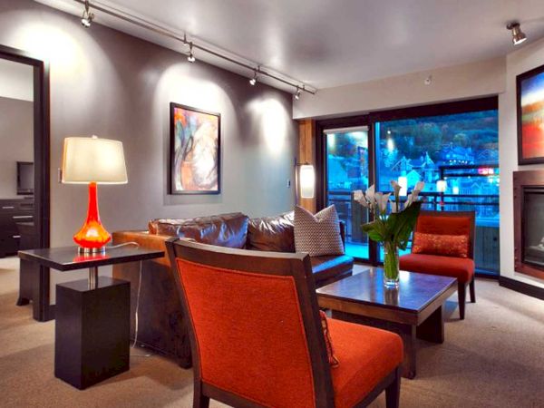A modern living room with orange chairs, a sofa, a fireplace, artwork, and a large window. Warm lighting enhances the cozy atmosphere.