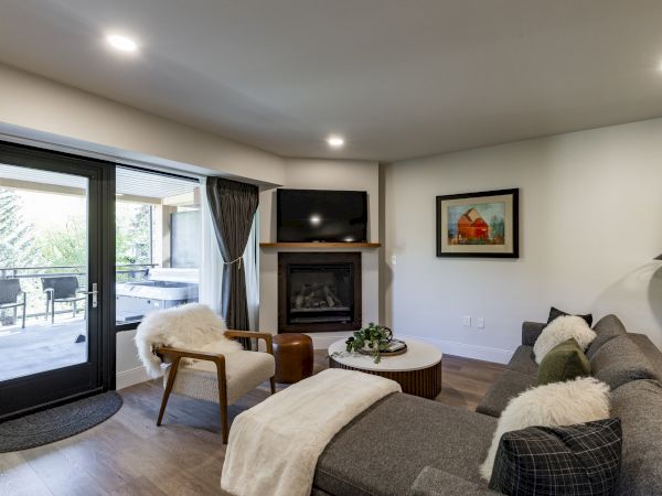 A cozy living room features a sectional sofa, fireplace, wall art, and a view of a patio with greenery outside through a glass door.