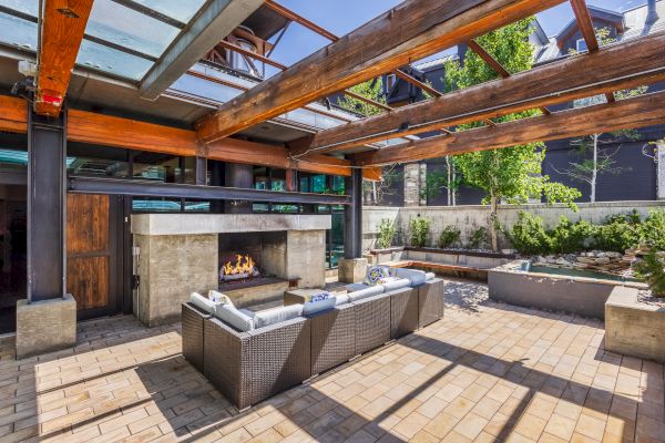 The image shows a modern outdoor patio with a sectional sofa, fireplace, wooden pergola, and garden area, creating a cozy and inviting space.