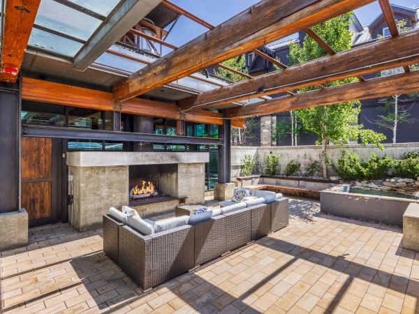The image shows a modern outdoor patio with a sectional sofa, fireplace, wooden pergola, and garden area, creating a cozy and inviting space.