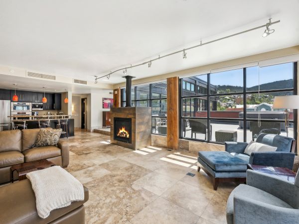 A modern living space with a fireplace, large windows, and comfy seating, connected to an open kitchen and outdoor view.