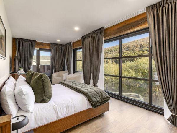 A modern bedroom with a large bed, pillows, side table, and floor-to-ceiling windows with curtains overlooking a scenic outdoor view.