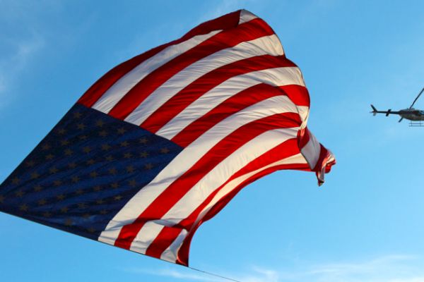 A large American flag waves in the sky as a helicopter flies nearby.