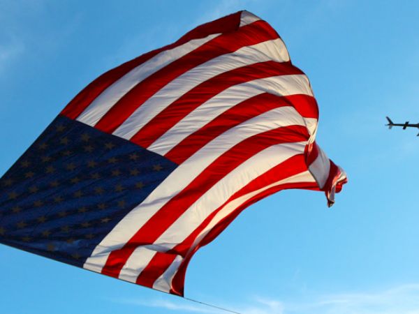 A large American flag waves in the sky as a helicopter flies nearby.