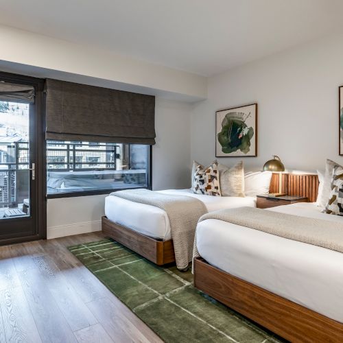 A modern hotel room with two twin beds, wooden cabinetry, large windows with blinds, framed artwork, and a green patterned rug.