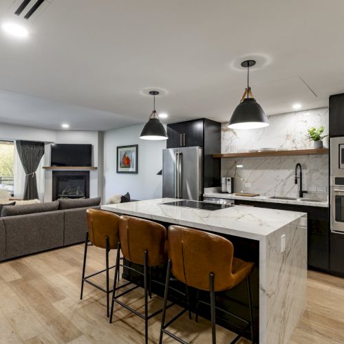 A modern kitchen and living area with an island, bar stools, stainless steel appliances, dark cabinets, and a cozy seating area.