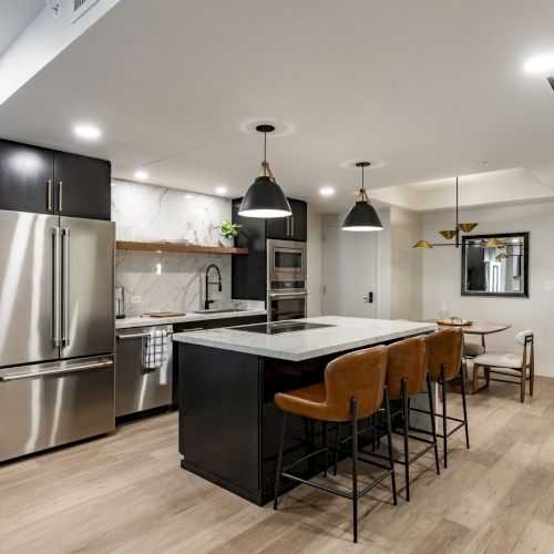 A modern kitchen with stainless steel appliances, a central island with seating, pendant lights, and wooden floors.