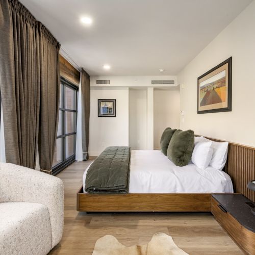 A modern bedroom with a large bed, dark curtains, and minimalistic decor, featuring wooden floors and a cozy chair in the corner.