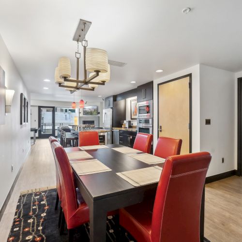 A modern dining area with a long table and red chairs, leading to a kitchen and a bedroom. Contemporary lighting and decor complete the look.