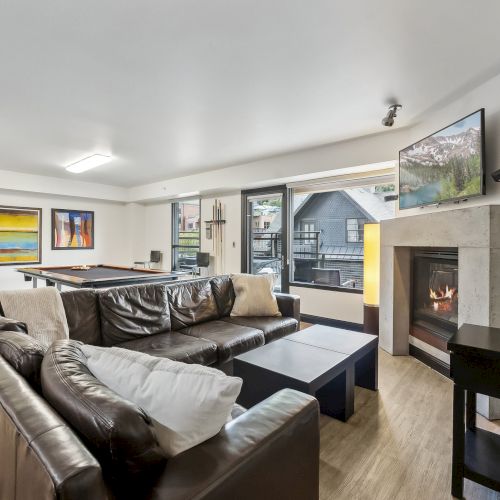 A modern living room features a brown sectional sofa, fireplace, wall art, TV, pool table, and a view through large windows.