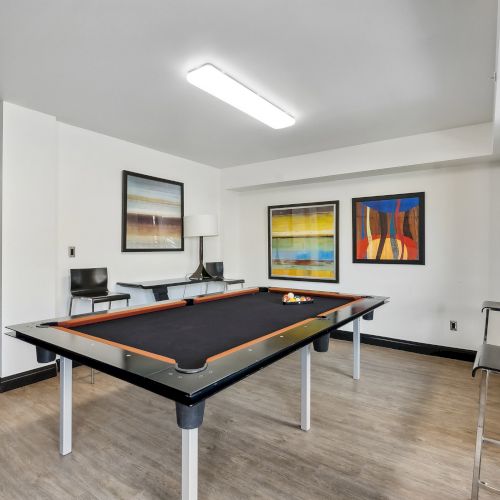 The image shows a modern room with a black pool table, colorful abstract art on the walls, and seating, under bright lighting.