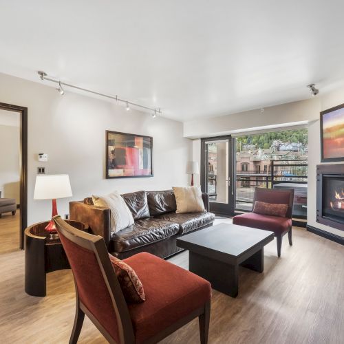 A modern living room features a sofa, chairs, a coffee table, a fireplace, and artwork on the walls. A balcony offers an outdoor view.