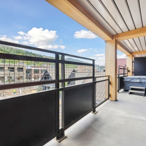 A balcony with railing overlooks buildings and hills; features a covered area with a hot tub and seating.